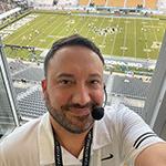 Jeff Sharon wears a headset microphone and smiles at the camera. He is in an announcer’s booth with the UCF football stadium below him.